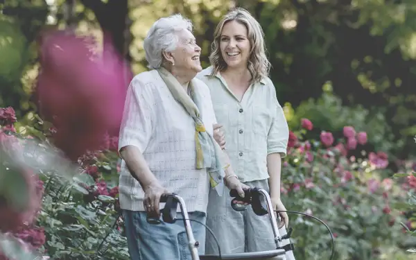 Eine Studentin der Sozialen Gerontologie begleitet eine ältere Frau mit einem Rollator auf einem Spaziergang durch einen Garten.