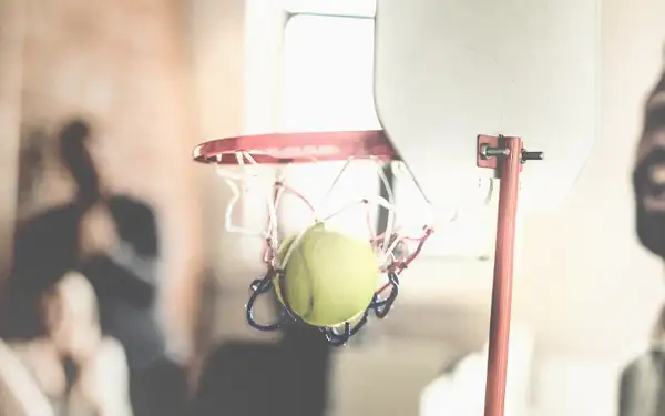 Sportmanager wirft Tennisball in kleinen Basketballkorb, im Hintergrund jubeln die Mitarbeiter. 