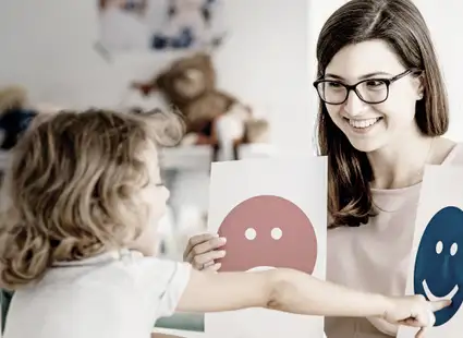 Eine Pädagogin zeigt einem Kleinkind auf spielerische Art und Weise Karten mit Smileys darauf. 