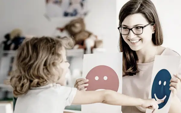 Eine Pädagogin zeigt einem Kleinkind auf spielerische Art und Weise Karten mit Smileys darauf. 