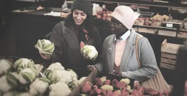 Zwei Frauen stehen vor einem Marktstand mit Obst und Gemüse und unterhalten sich über ihre Ausbildung zum Ernährungscoach. 