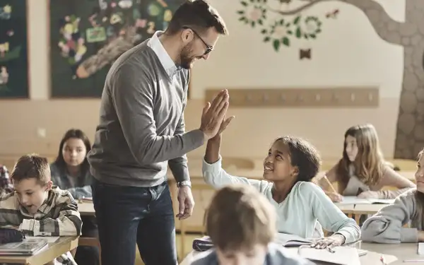 Ein Grundschulpädagoge gibt einer Schülerin in einem Klassenzimmer ein High-Five. Die Schülerinnen und Schüler arbeiten an ihren Aufgaben und sind aufmerksam.