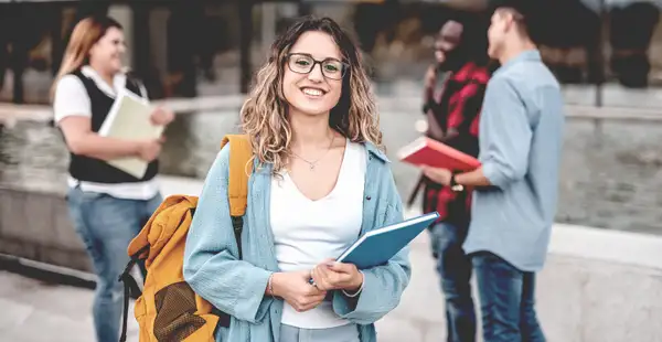 Eine Studentin steht auf dem Unigelände mit einem Buch im Arm und lächelt.