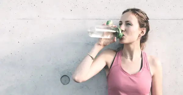 Junge Sportlerin trink an eine Wand gelehnt lächelnd Wasser.