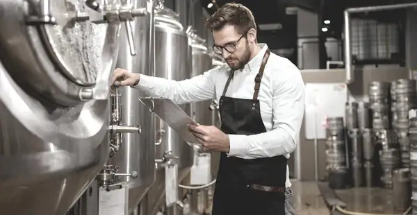 Ein Bierbrauer, der Brauwesen und Getränketechnologie studiert hat, steht vor einem Braukessel und nimmt Einstellungen vor. In der Hand hält er ein Klemmbrett und macht sich Notizen.