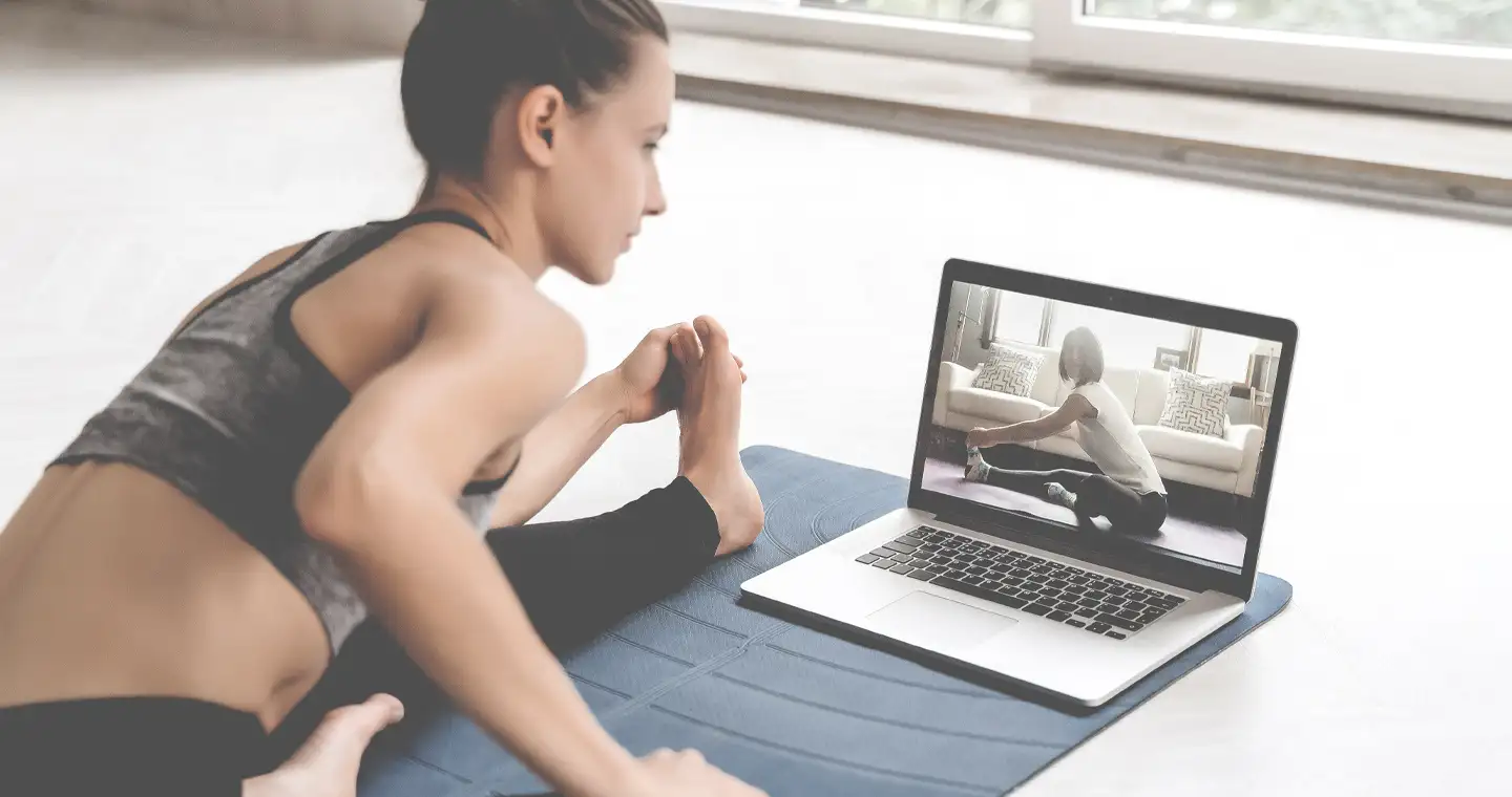 Eine Frau sitzt in Sportkleidung auf einer Sportmatte und macht Dehnübungen vor dem Laptop.