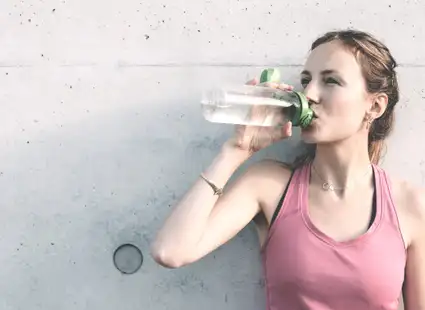 Junge Sportlerin trink an eine Wand gelehnt lächelnd Wasser.