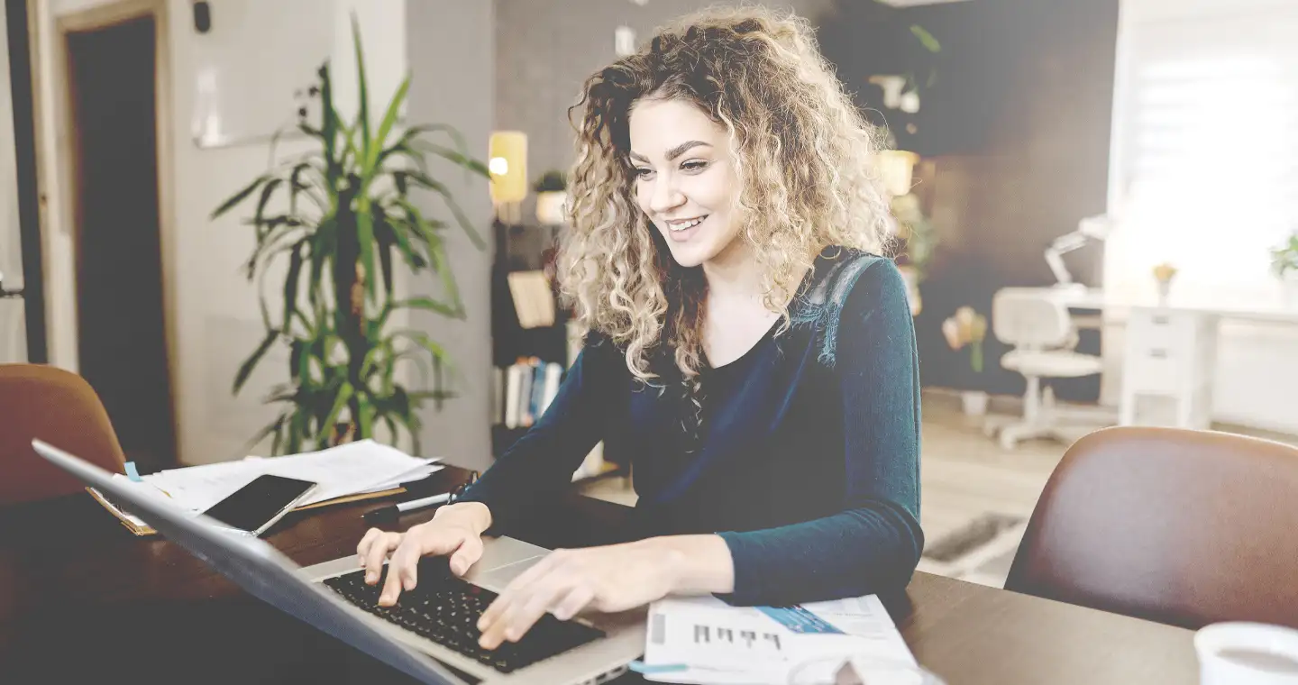 Eine junge Frau mit Locken sitzt lächelnd am Laptop und bildet sich weiter.