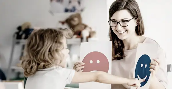 Eine Pädagogin zeigt einem Kleinkind auf spielerische Art und Weise Karten mit Smileys darauf. 