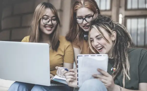 Drei Studentinnen, die Gender Studies studieren, arbeiten gemeinsam an einem Projekt. Sie lachen und diskutieren, während sie einen Laptop und Notizbücher verwenden.