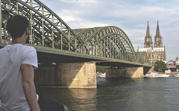 Ein Student sitzt am Rhein und blickt auf die Hohenzollernbrücke und den Kölner Dom.