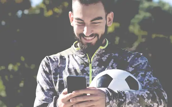 Ein Sportdirektor in Fußball hat einen Fußball unter den Arm geklemmt und tippt an seinem Smartphone.