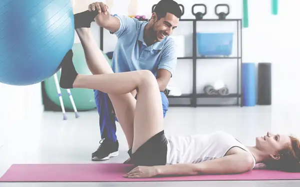 Ein Sporttherapeut unterstützt eine Patientin bei einer Übung auf einer Matte.