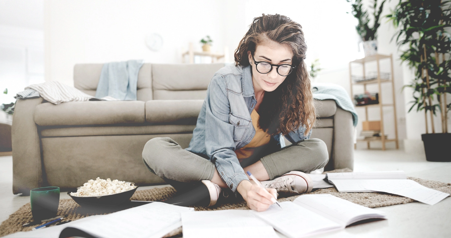 Eine junge Frau sitzt vor ihrer Couch auf dem Boden und macht Notizen in ihren Unterlagen für die Studienfinanzierung.