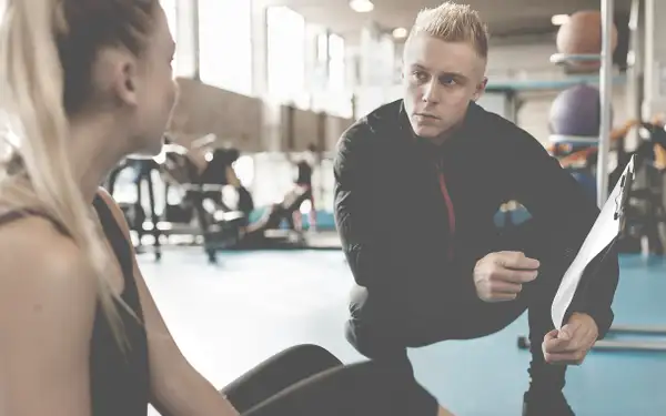 Sportpsychologe kniet neben Sportlerin im Fitnessraum und erklärt ihr etwas