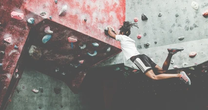 Ein sportlicher junger Mann in lässigen Sportklamotten lässt beim Bouldern an der Kletterwand seine Beine baumeln.