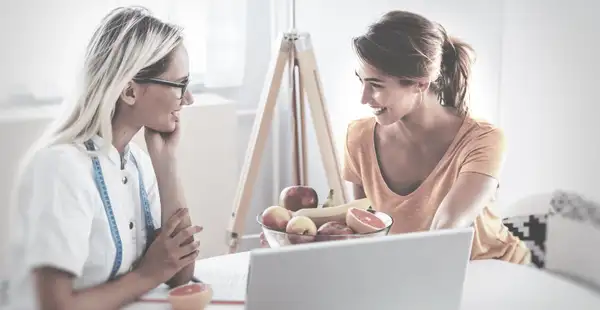 Eine Ernährungsberaterin sitzt mit ihrer Klientin in einem Besprechungszimmer, sie unterhalten sich mit einem Lächeln auf den Lippen, vor ihnen steht eine Schale mit Obst.