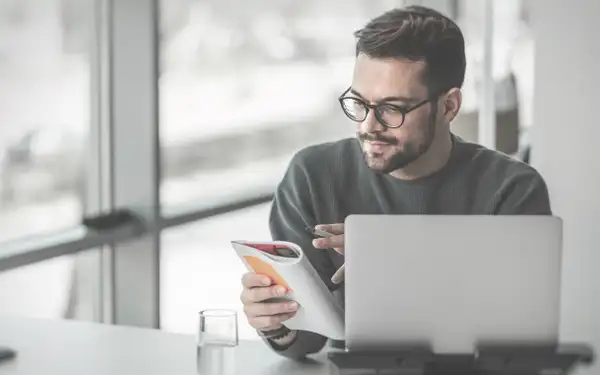 Ein Online Marketer sitzt im Büro vor seinem weißen Laptop und hält einen Schreibblock in der einen und einen Kugelschreiber in der anderen Hand, als würde er gleich etwas notieren.