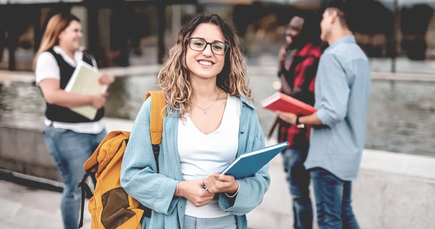 Eine Studentin hält ein Buch im Arm und lächelt in die Kamera.