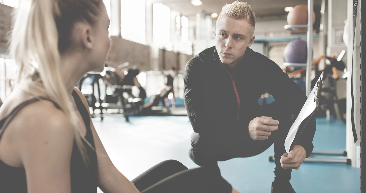 Sportpsychologe kniet neben Sportlerin in Fitnessstudio und erklärt ihr etwas