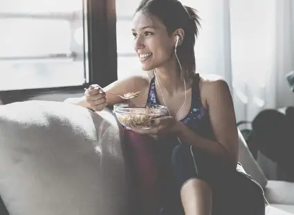 Sportlerin sitzt in Sportbekleidung auf dem Sofa und isst Müsli