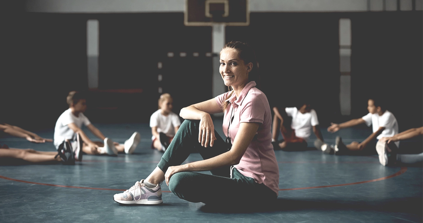 Junge Sportpädagogin sitzt gemeinsam mit ihren Schüler*innen auf dem Hallenboden.