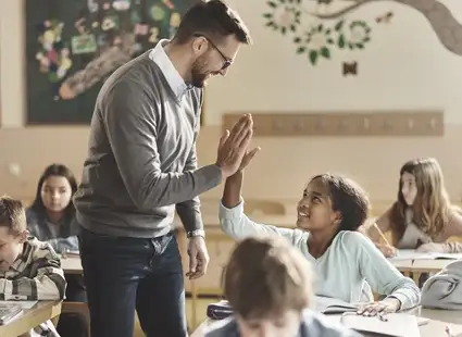 Ein Grundschulpädagoge gibt einer Schülerin in einem Klassenzimmer ein High-Five. Die Schülerinnen und Schüler arbeiten an ihren Aufgaben und sind aufmerksam.