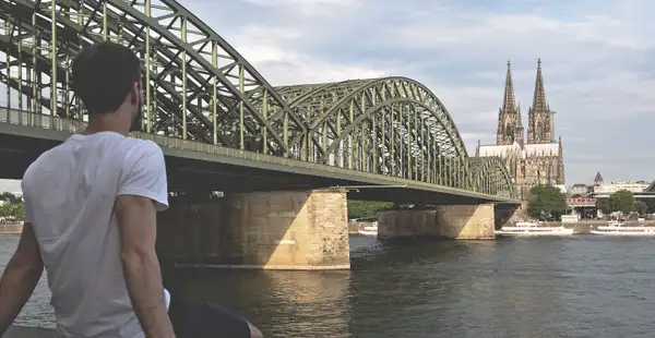 Ein Student sitzt am Rhein und blickt auf die Hohenzollernbrücke und den Kölner Dom.