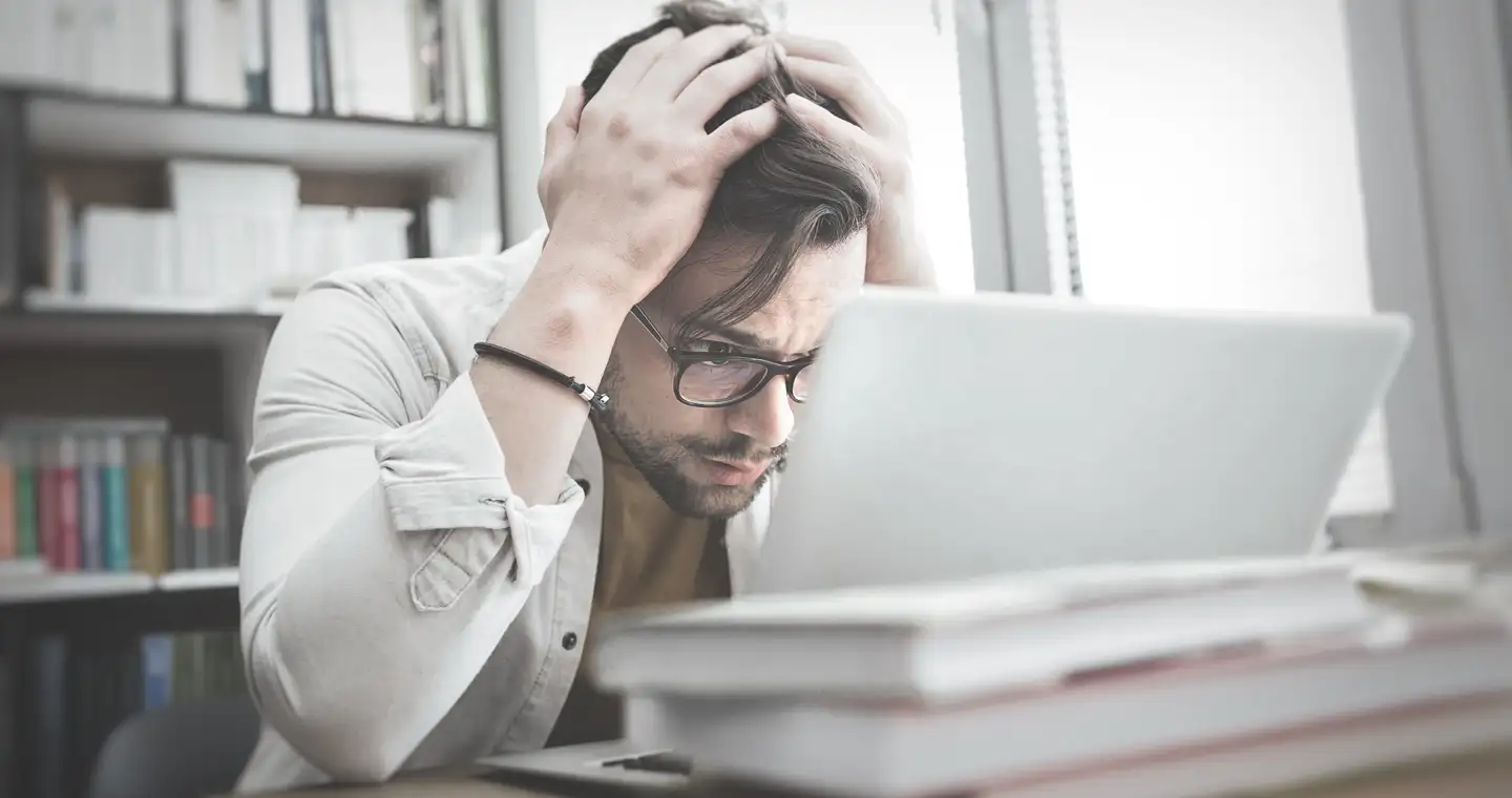 Ein Wirtschaftspsychologie Student hält sich beim Lernen den Kopf und schaut überfordert auf seinen Laptop.