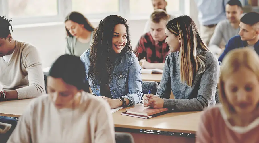 Zwei Studentinnen sitzen in einem vollen Seminarraum am Tisch und unterhalten sich lächelnd