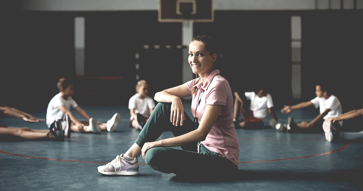 Sportlehrerin sitzt auf dem Hallenboden und zeigt ihren Schülern Aufwärmübungen