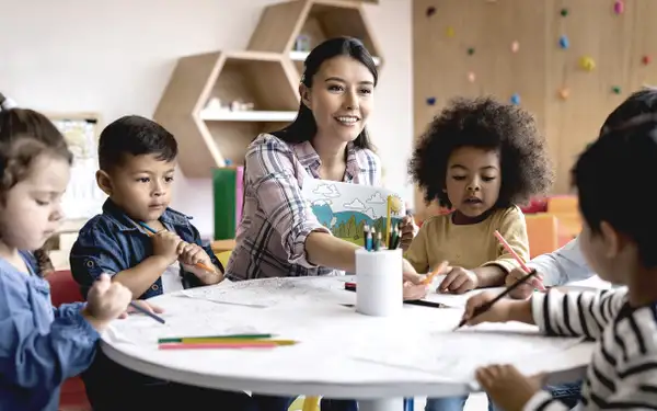 Eine Sozialpädagogin sitzt an einem runden Tisch und malt zusammen mit einer Gruppe von Kindern.