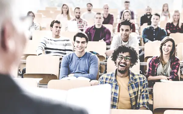 In einem Hörsaal sitzen lächelnde Studierende und hören einem Professor zu. 