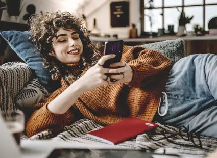 Eine junge Medienpsychologie Studentin liegt mit einem Buch und ihrem Handy in den Händen auf der Couch.