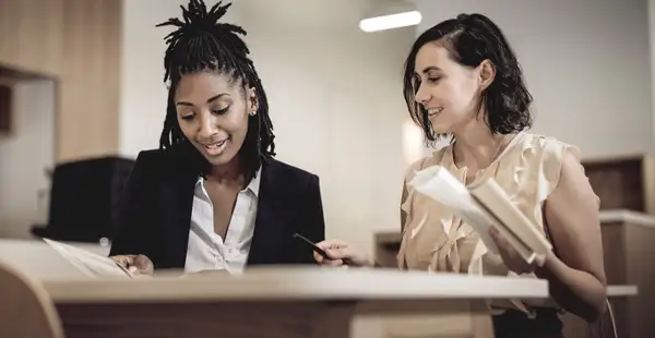 Zwei Studentinnen, die dual Sozialmanagement studieren, arbeiten lächelnd gemeinsam an Dokumenten in einem modernen Büro.