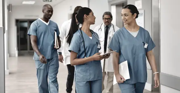 Eine Gruppe von medizinischem Personal, darunter zwei Frauen in blauen Pflegeuniformen, geht durch einen Krankenhausflur und unterhält sich.