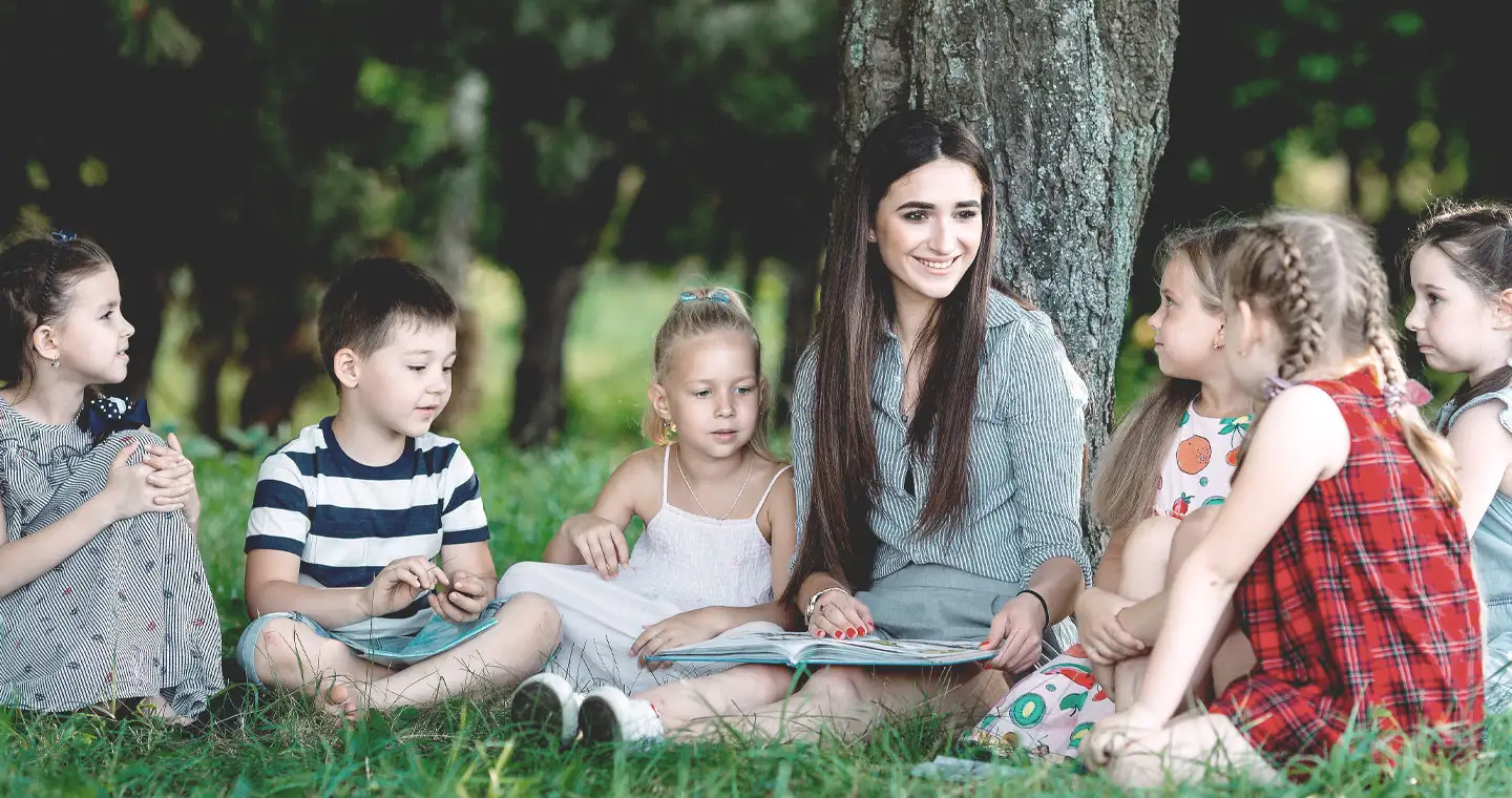 Eine Erzieherin sitzt mit einer Gruppe von Kindern auf einer Wiese und liest ihnen vor. 