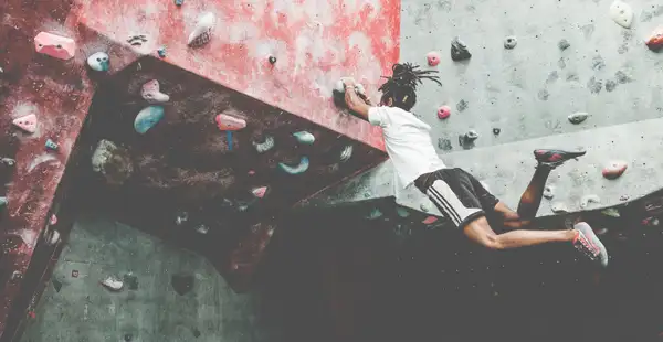 Ein sportlicher junger Mann in lässigen Sportklamotten lässt beim Bouldern an der Kletterwand seine Beine baumeln.