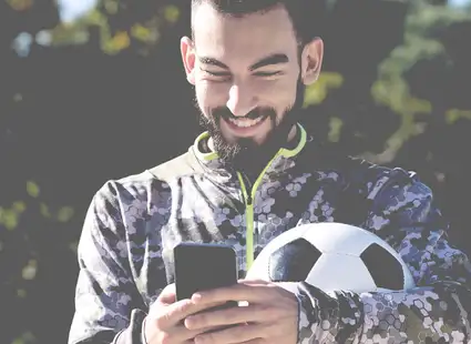 Ein Sportdirektor in Fußball hat einen Fußball unter den Arm geklemmt und tippt an seinem Smartphone.