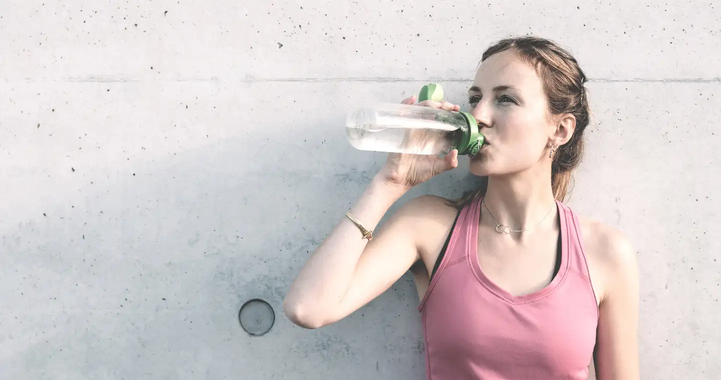 Junge Sportlerin trinkt an eine Betonwand gelehnt Wasser aus einer Trinkflasche.