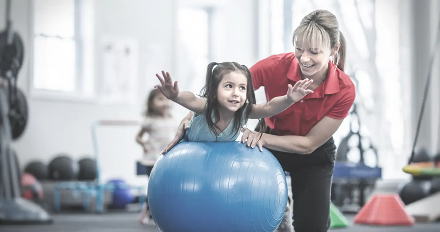 Eine Sporttherapeutin führt eine Bewegungstherapie mit einem Mädchen auf einem Gymnastikball durch. 