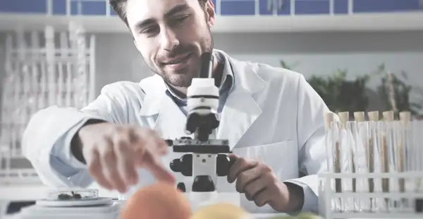 Ein Ernährungswissenschaftler greift nach einer Orange, in der Hand hält er ein Mikroskop. 