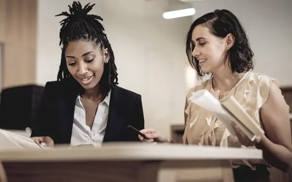 Zwei Studentinnen, die dual Sozialmanagement studieren, arbeiten lächelnd gemeinsam an Dokumenten in einem modernen Büro.