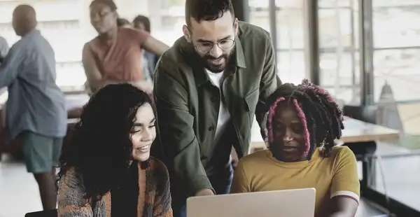 Ein Sozialarbeiter hilft zwei jungen Frauen an einem Laptop. Alle drei lächeln und wirken konzentriert.