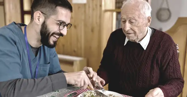 Ein Pfleger, der eine Weiterbildung in der Pflege absolviert, hilft einem älteren Mann beim Essen. Beide lachen und wirken glücklich.