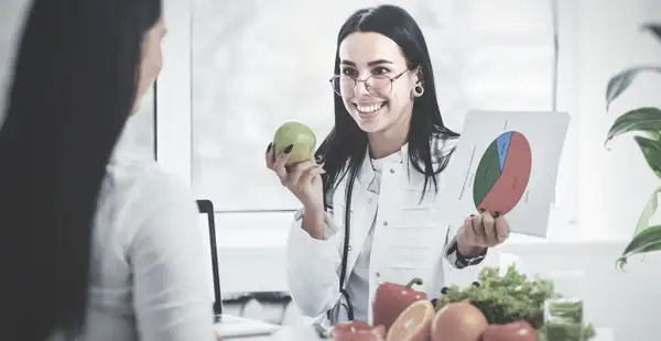 Eine Frau, die eine Weiterbildung zur Gesundheitsberaterin absolviert hat, unterhält sich mit ihrer Klientin, während sie einen Apfel und ein Blatt mit einem Diagramm in den Händen hält. 