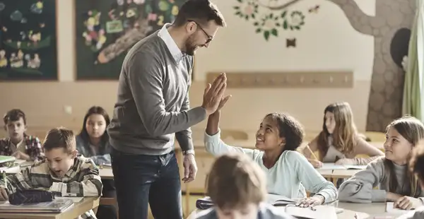 Ein Grundschulpädagoge gibt einer Schülerin in einem Klassenzimmer ein High-Five. Die Schülerinnen und Schüler arbeiten an ihren Aufgaben und sind aufmerksam.