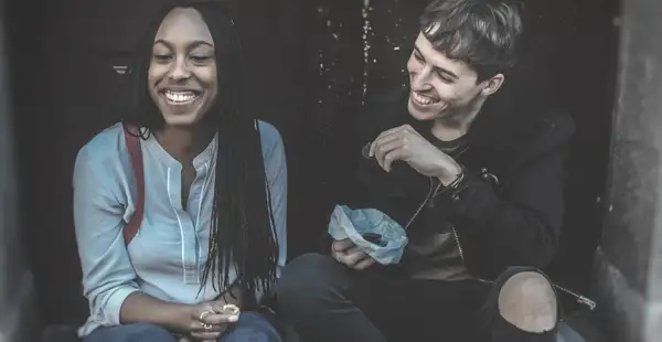 Auf einer Treppe sitzen ein junger Mann und eine junge Frau, die beide Soziale Arbeit berufsbegleitend studieren. Sie lächeln und der Mann hält eine Tüte mit Essen in der Hand.