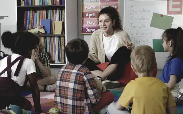 Eine junge Frau, die ein Fernstudium in Kindheitspädagogik absolviert, sitzt auf dem Boden eines Klassenzimmers und unterrichtet eine Gruppe von Kindern.