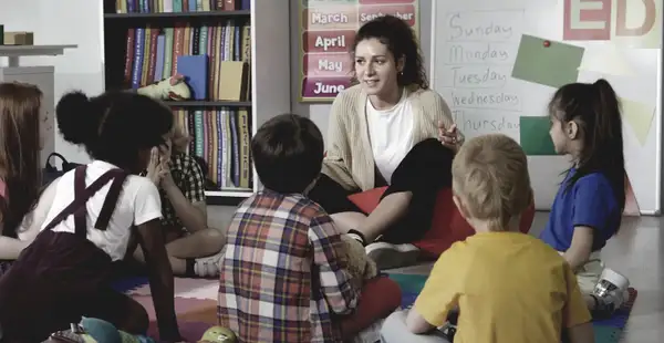 Eine junge Frau, die ein Fernstudium in Kindheitspädagogik absolviert, sitzt auf dem Boden eines Klassenzimmers und unterrichtet eine Gruppe von Kindern.